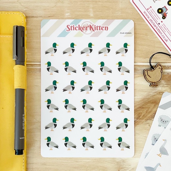 Duck stickers by StickerKitten on a wooden board with the edge of a planner and pen visible to the left and a chicken planner charm and sheep stickers on the right
