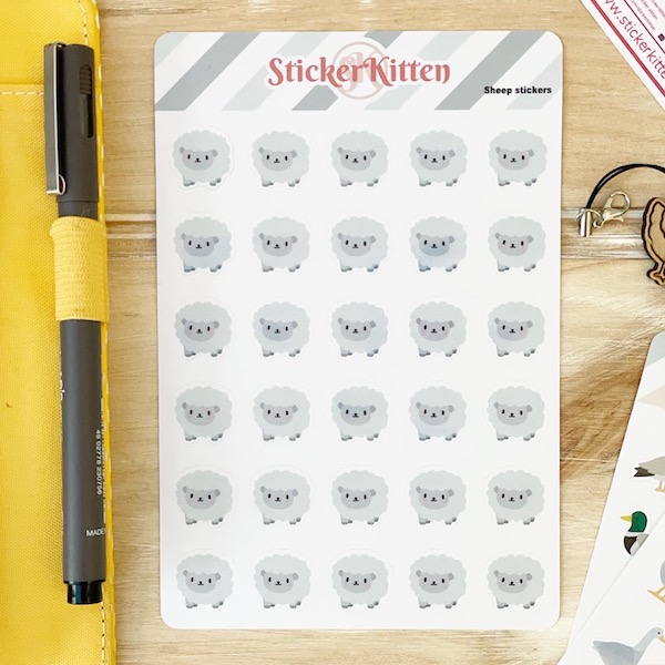 A sheet of cute sheep stickers on a wooden board next to a planner with a pen