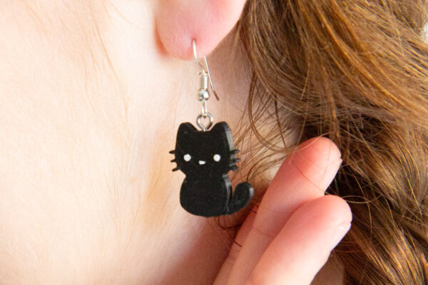 StickerKitten black cat dangly earring worn in ear. Cute black cat silhouette with white painted face.